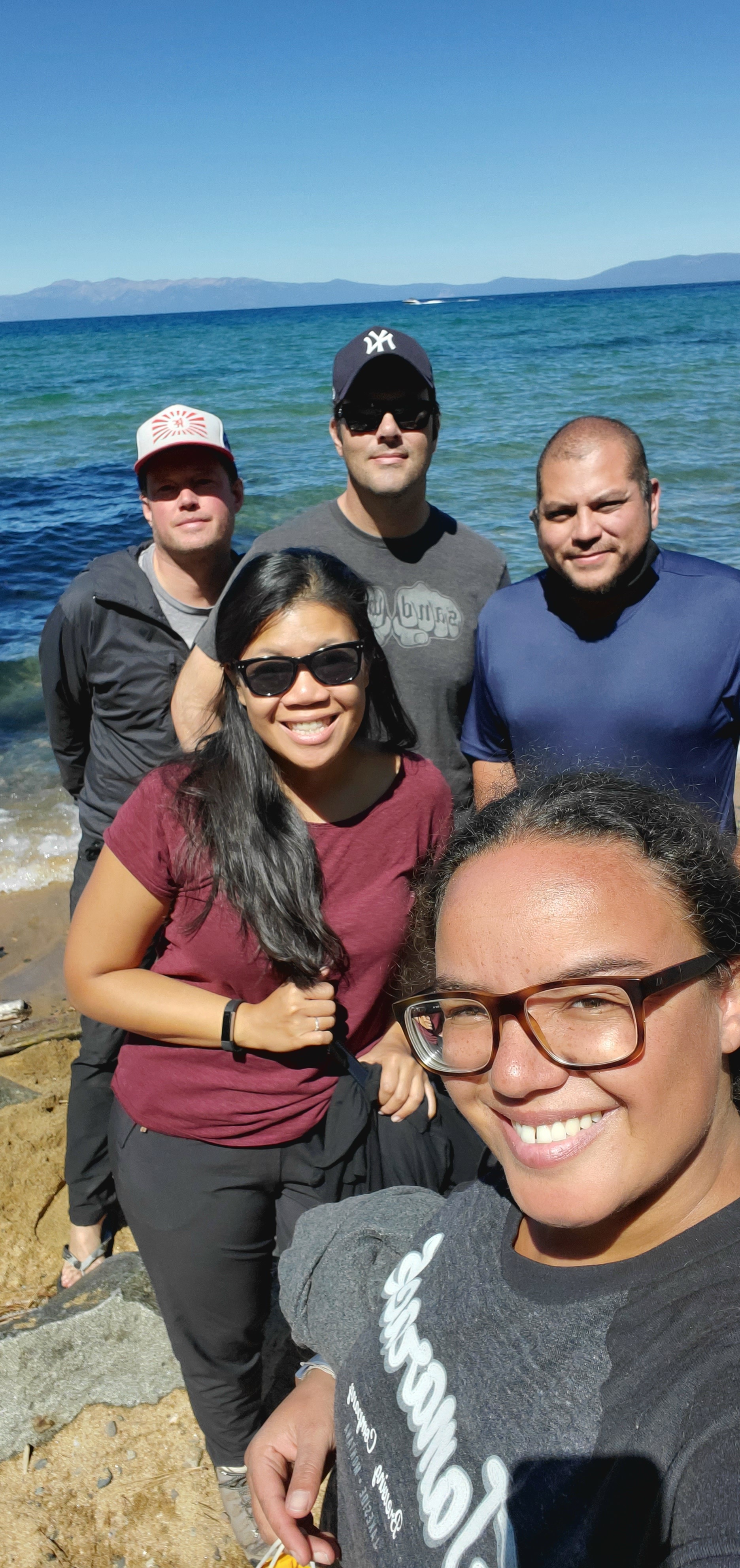 Five friends standing on a beach with the water and mountains in the background.